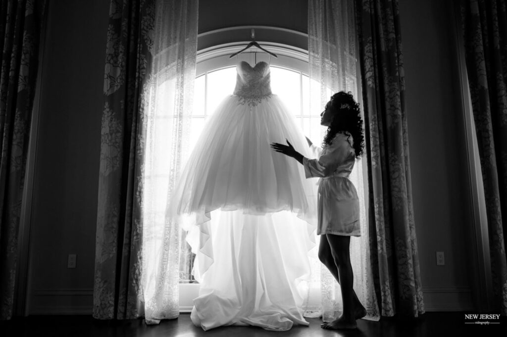 Bride in a robe admiring her wedding dress hanging in front of a large window